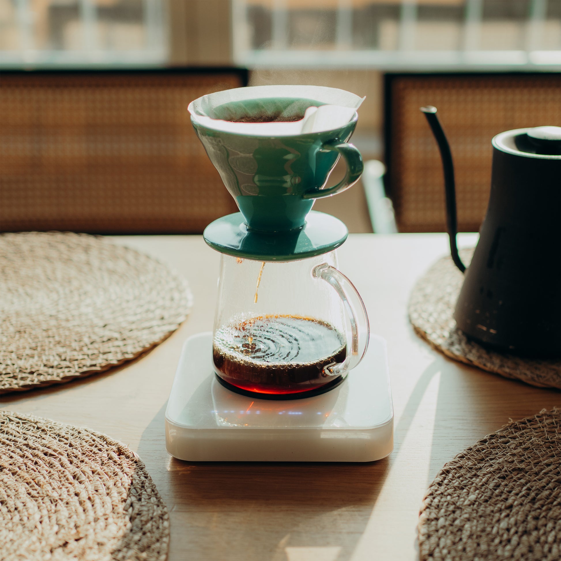 Teal coffee dripper with glass carafe on a white surface, surrounded by woven coasters.